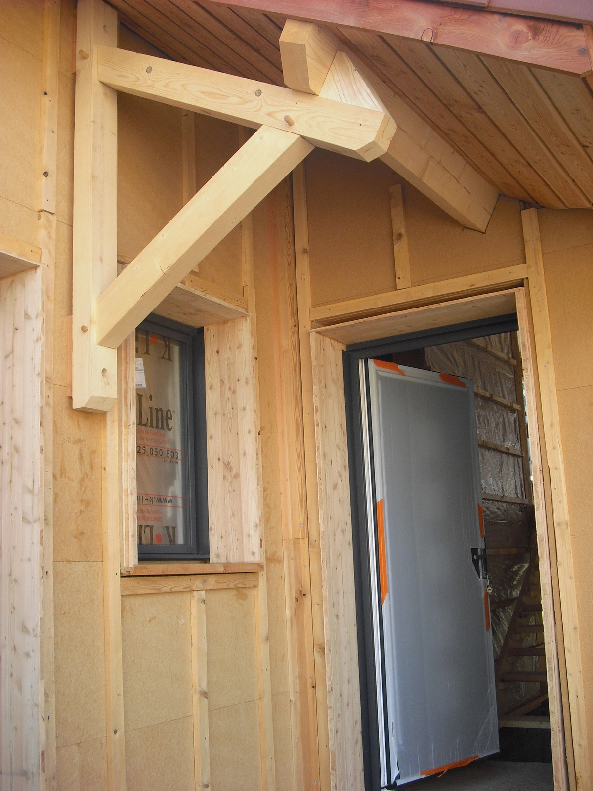 Détail d'une charpente en bois d'une maison en construction, montrant un auvent au-dessus d'une porte d'entrée et une fenêtre. La structure en bois brut est visible, avec des poutres apparentes et un bardage non fini.