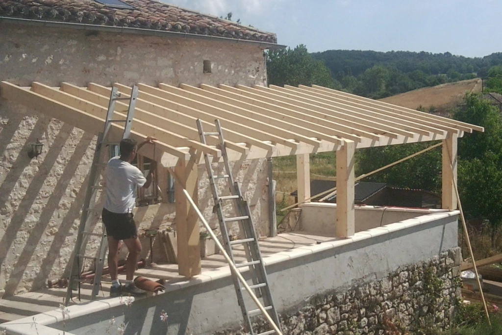 Construction d'une extension en bois sur une maison en pierre, montrant un charpentier travaillant sur la structure du toit. La scène se déroule dans un environnement rural avec des collines boisées en arrière-plan.