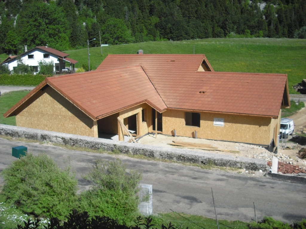 Maison en construction avec charpente et toiture en tuiles rouges, entourée de verdure et de forêts